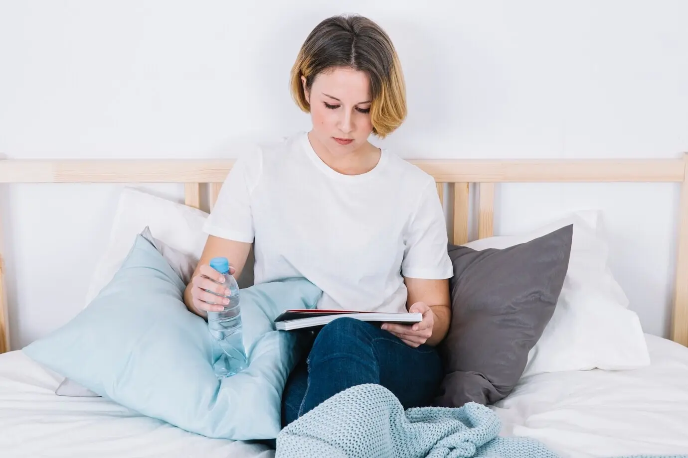 A woman reads on a bed with a water bottle.