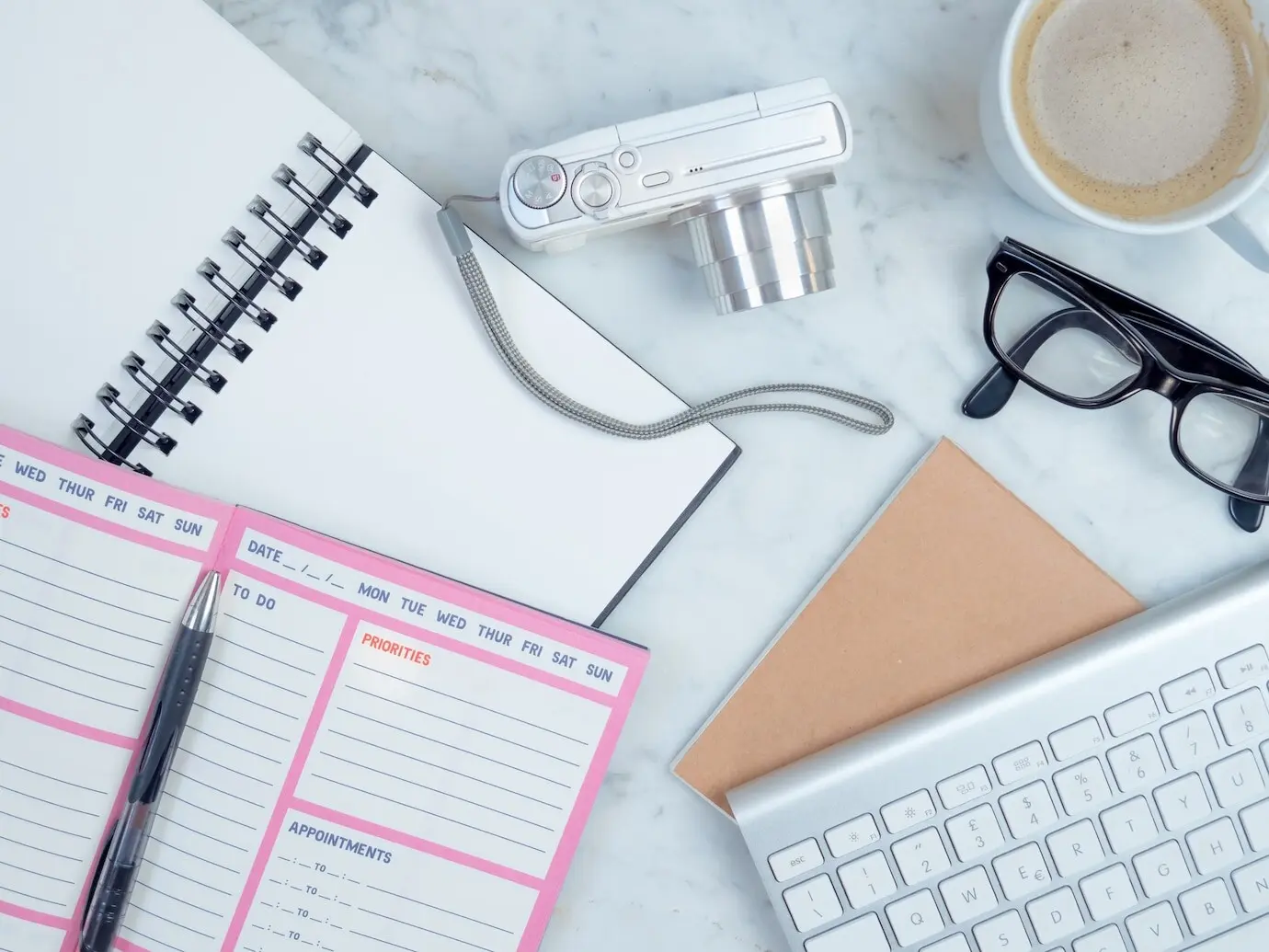 Close-up, top-down view of the notebook, planner, eyeglasses, keyboard, coffee, and camera on the bed.