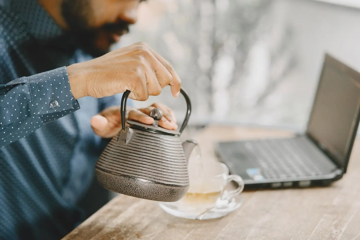 An African-American man is working on a laptop and writing in a notebook. A bearded man is sitting in a cafe and pouring tea.