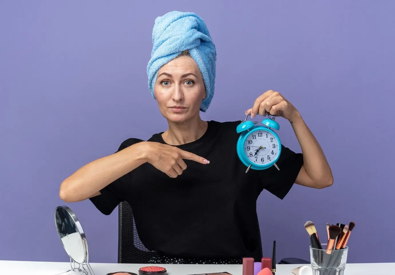 A happy young, beautiful girl sits at a table with makeup tools, drying her hair with a towel, holding an alarm clock and pointing at it, isolated against a blue wall.